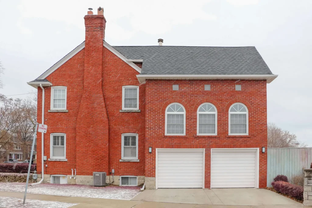 A large, two-story red brick house with a prominent chimney and two white garage doors, a perfect candidate for considering painting brick exterior