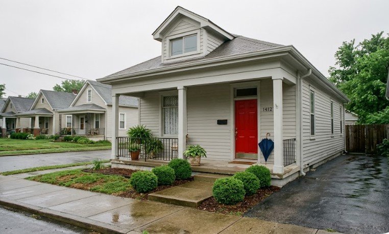 Light gray small house with red front door, white porch, and wet sidewalk after rain in Louisville KY